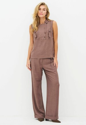 Femme aux longs cheveux blonds portant un haut marron sans manches avec bretelles et un pantalon large assorti, debout devant un fond blanc.