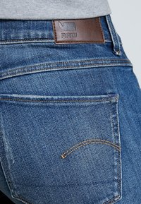 Blauwe denim jeans met een bruine leren patch met het "RAW" logo, zichtbare stiksels en een standaard ontwerp van de achterzak.