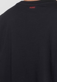 Camiseta negra hecha de algodón suave. Presenta un cuello redondo y un pequeño logo rojo en la parte posterior cerca del cuello. Diseño simple y minimalista.