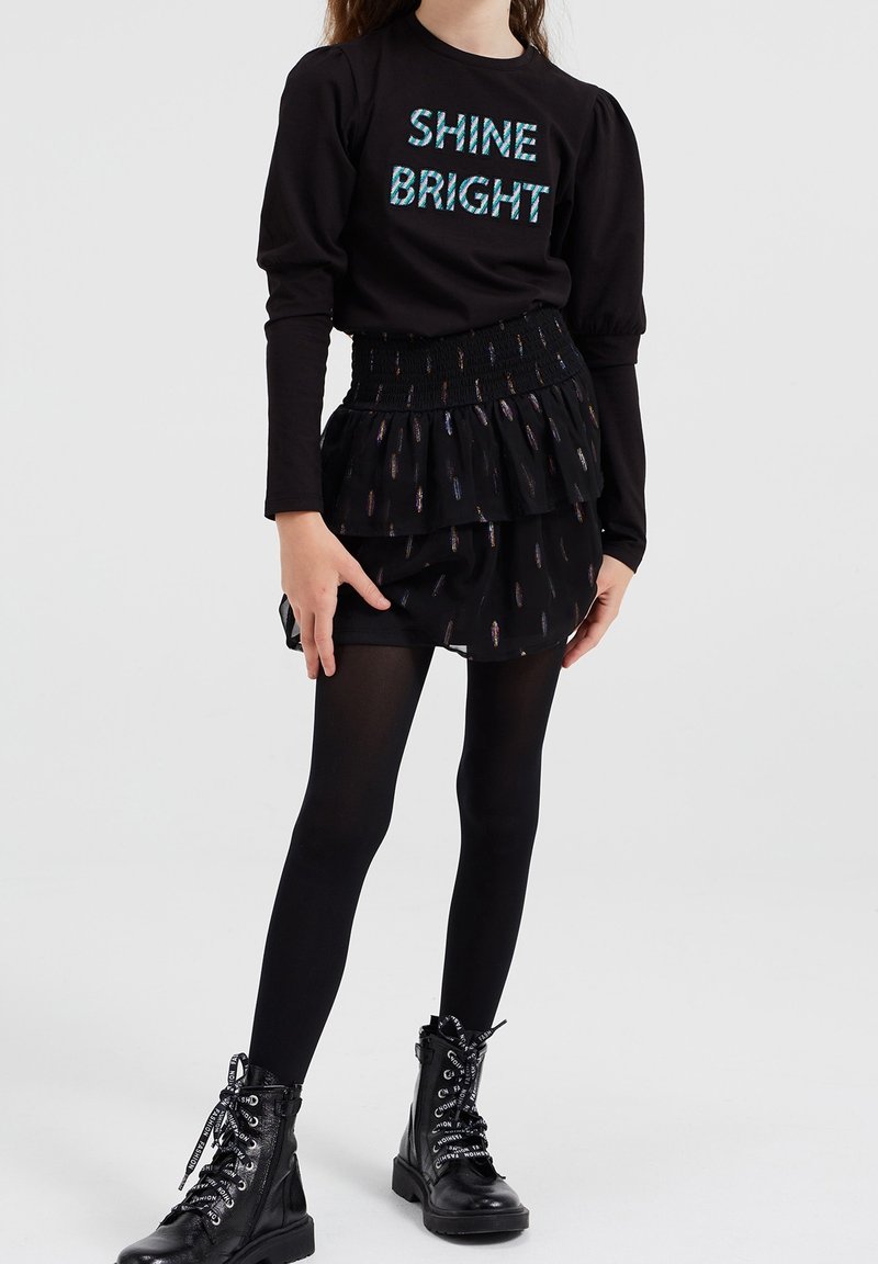 Fille portant un haut noir à manches longues avec le texte "SHINE BRIGHT", une jupe noire superposée avec des détails brillants, des collants noirs et des bottes de combat noires.