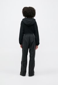 Άτομο με σγουρά μαλλιά που φοράει μαύρο φούτερ με κουκούλα και μαύρο παντελόνι με λευκές πλευρικές ρίγες, στέκεται με την πλάτη στο λευκό φόντο.