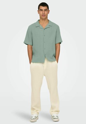 Green short-sleeve button-up shirt in textured fabric, paired with loose-fitting cream textured pants and white sneakers.