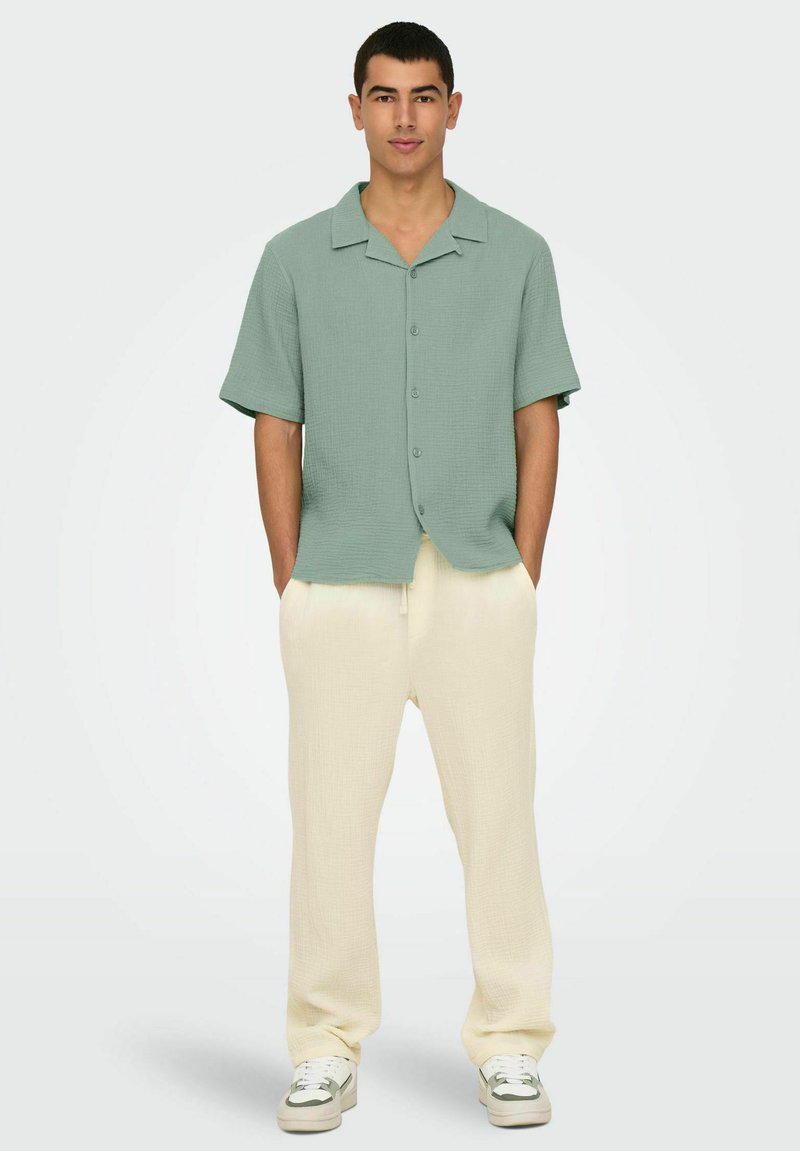 Chemise à manches courtes verte à boutons en tissu texturé, associée à un pantalon ample crème en tissu texturé et des baskets blanches.