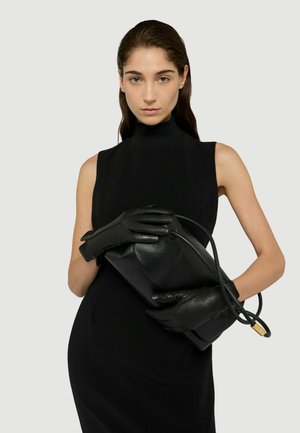 Schwarze Lederclutch, gehalten von behandschuhten Händen; glatte Textur, rechteckige Form, minimalistisches Design. Model in einem ärmellosen schwarzen Kleid.