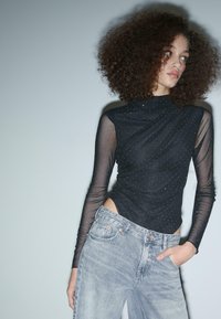 Schwarzer Bodysuit mit transparenten, gepunkteten langen Ärmeln und einem drapierten Design, kombiniert mit hellblauen, hochgeschnittenen Jeans mit klassischen Knopfdetails.
