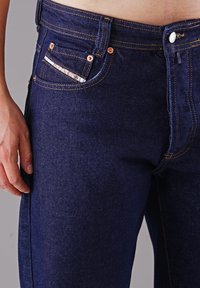 Dunkelblaue Jeans aus Denim mit orangefarbenen Nähten, kupfernen Nieten und einem kleinen gemusterten Taschendetail. Nahaufnahme der Taille und der Hand.