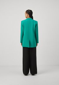 Grüner Blazer mit langen Ärmeln, tailliertem Design und Rückenventil, kombiniert mit weiten schwarzen Hosen. Glattes Material und klare Linien.