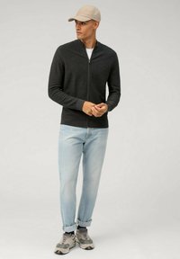 Donkergrijze ritsvest met ribbelstructuur, gedragen over een wit shirt, in combinatie met lichtblauwe jeans en grijze sneakers. Beige pet op het hoofd.