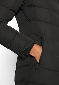 Černá péřová bunda s prošívaným designem, zipovou kapsou a žebrovanými manžetami. Hladká, lehká tkanina s matným povrchem.