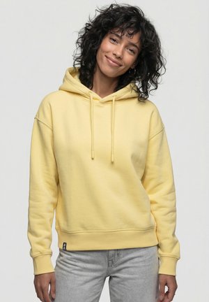 Frau mit lockigem dunklem Haar, die einen gelben Hoodie und hellgraue Jeans trägt und sanft lächelt vor einem schlichten hellen Hintergrund.