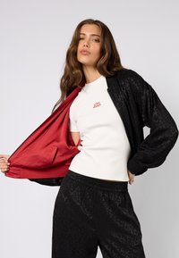 Czarna kurtka bomber z podszewką w panterkę, czerwoną wyściółką, noszona rozpięta na białą dopasowaną koszulkę z napisem "LOVE", zestawiona z czarnymi spodniami.