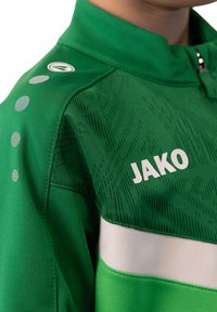 Groene sportjack met een ritssluiting bij de kraag, voorzien van textuurpatronen, witte strepen en het opdruklogo "JAKO" op de borst.