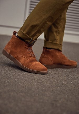 Brune ruskindsankelstøvler med rund tå, snørebåndsdesign og en struktureret gummisål. Parret med optrukne olivengrønne bukser.