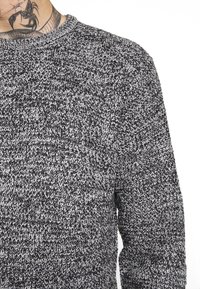 Svetr s texturovaným úpletem v šedých a černých tónech, s kulatým výstřihem a dlouhými rukávy. Materiál působí měkce a teple.