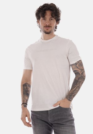 Uomo con capelli ricci e barba che indossa una maglietta bianca e jeans grigi, mostrando tatuaggi dettagliati neri e grigi su entrambi gli avambracci.