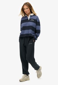 Polo de manga larga a rayas azules con cuello blanco, combinado con pantalones de chándal negros con un logo y zapatillas deportivas beige. Estilo casual, ajuste relajado.