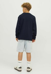 Berniukas su garbanotais plaukais vilkintis tamsiai mėlyną ilgųjų rankovių marškinį, šviesius džinsinius šortus, firmines baltas kojines ir baltus sportinius batus, stovintis atsisukęs atgal.