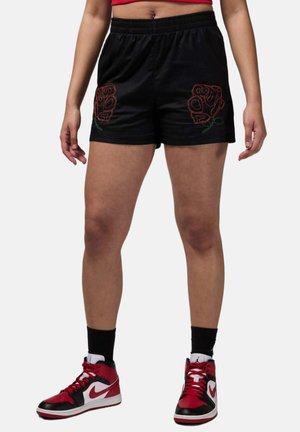 Henkilö, joka on pukeutunut mustiin shortseihin, joissa on punaisen ruusun kirjailu kummassakin lahkeessa, mustiin sukkiin sekä punaisiin, mustiin ja valkoisiin Nike-tennareihin, seisomassa jalat erillään.