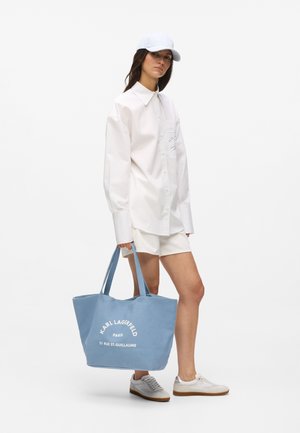 Kvinne iført hvit, oversized skjorte, hvite shorts, hvit caps og joggesko, som holder en stor blå Karl Lagerfeld handlenett.