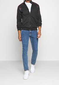 Giacca nera con zip e motivo a quadri sulle spalle, abbinata a jeans blu attillati e sneakers bianche. Tessuto morbido, vestibilità casual.
