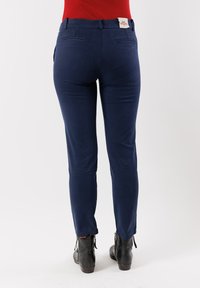 Persona che indossa pantaloni chino blu navy aderenti e stivali neri alla caviglia, in piedi contro uno sfondo bianco semplice, vista posteriore.