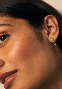 Nahaufnahme eines Frauen Gesichts mit glatter Haut, natürlichen Sommersprossen und einem goldenen Creolen-Ohrring mit einem diamantähnlichen Anhänger.