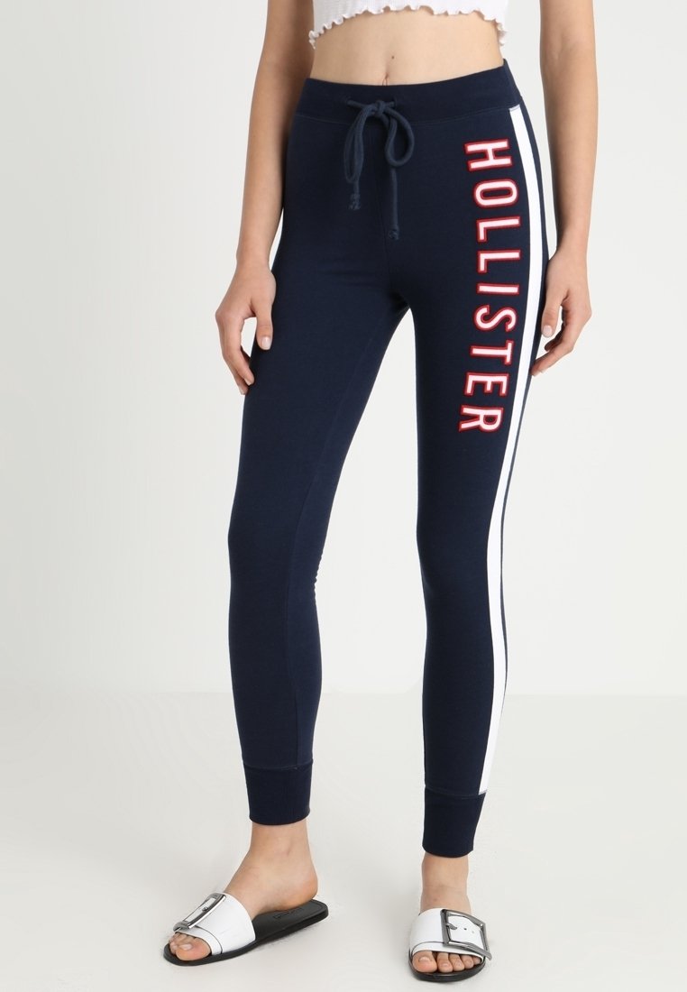 Leggings azul marino con franjas laterales blancas, con el texto rojo "HOLLISTER" en la pierna izquierda. Fabricados con una tela elástica y con una cinturilla elástica.