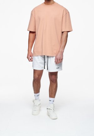 Lichtroze katoenen t-shirt met korte mouwen, gecombineerd met witte sportshorts met zwarte trekkoorden en merkopdruk. Witte sneakers en korte sokken.