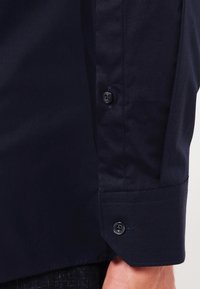 Camisa azul marino con una textura suave, que presenta un puño ajustado, dos botones negros y un detalle de tapeta de manga con un solo botón.