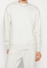 Man i ljusbeige matchande sweatshirt och träningsoverallsbyxor med subtilt Jumpman-logga, silverhalsband, står med avslappnad hand.