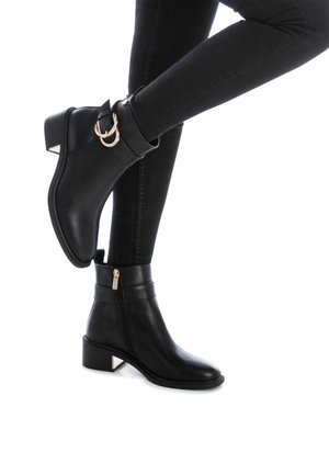 Botas de cuero negro hasta el tobillo con cierre de cremallera, acentos de hebilla dorada y un tacón bajo de bloque con punta plateada. Textura suave.