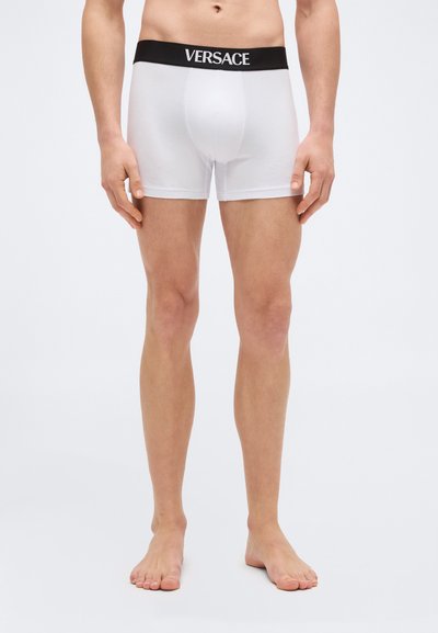 Ανδρικός κορμός και πόδια που φορούν λευκά ανδρικά εσώρουχα Versace με μαύρη ζώνη στη μέση, σε απλό φόντο.