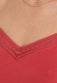 Débardeur rouge à décolleté en V avec une délicate bordure en dentelle le long du col. Texture lisse et design ajusté.