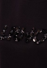 Tessuto nero con testo parzialmente visibile decorato con piccoli paillettes neri e argentati che creano un effetto scintillante.