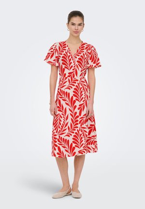 Femme debout portant une robe portefeuille mi-longue à motifs de feuilles rouges sur fond blanc, et des chaussures plates tissées beiges.
