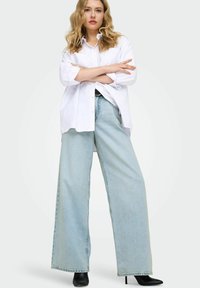 Jean en denim bleu clair à jambes larges associé à une chemise blanche oversize avec col classique et manches retroussées.