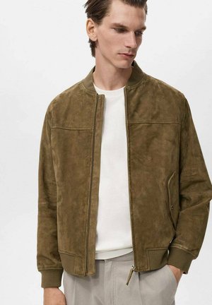 Veste bomber en suède marron avec une fermeture éclair à l'avant, des poignets côtelés et des poches latérales. Portée par-dessus une chemise blanche, présentant un éclat subtil et une texture lisse.