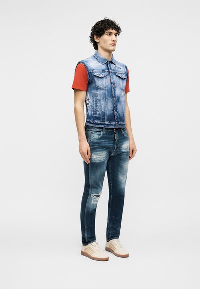 Νεαρός άνδρας στέκεται, φοράει μπλε τζιν γιλέκο χωρίς μανίκια πάνω από κόκκινο t-shirt, φθαρμένα μπλε τζιν και μπεζ αθλητικά παπούτσια με καουτσούκ σόλες.