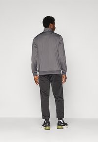 Mand, der vender væk, iført en grå zip-jacket, sorte joggerbukser, hvide sokker og sorte Puma sneakers med grønne såler.