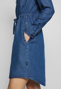 Närbild av en person som bär en blå denimklänning med långa ärmar, elastiskt midjeband, sidofickor och ett tygbälte.