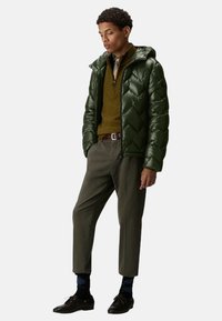 Giacca imbottita verde scuro con motivo a chevron, chiusura con zip e cappuccio indossata sopra un maglione a coste color senape, abbinata a pantaloni olive e scarpe scure.