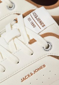Weiße Sneaker mit einem glatten Lederobermaterial, strukturierten braunen Akzenten, flachen weißen Schnürsenkeln und einem Logo-Patch auf der Zunge. Mit durchbrochene Details versehen.