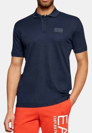 Uomo che indossa una polo blu navy a maniche corte con logo EA7 e pantaloncini arancioni brillanti con marchio Emporio Armani.