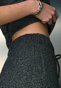 Zwarte top met glitters en bijpassende rok met elastische tailleband. De stof heeft een metallic glans en een gladde textuur. Zilveren armband zichtbaar.