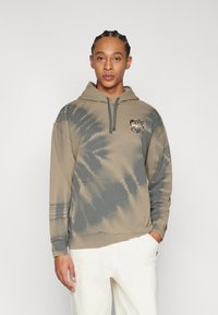 Hoodie tie-dye beige et gris avec une poche avant et un capuchon à lien de serrage. Présente un logo sur la poitrine et des poignets et un ourlet côtelés.