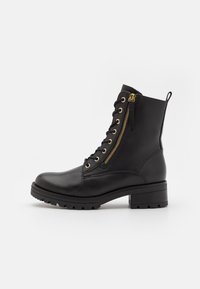 Zwarte leren enkelboot met gouden hardware, veters aan de voorkant, zijrits, en een dikke, gestructureerde zool. Heeft een blokhak en een lus aan de achterkant.