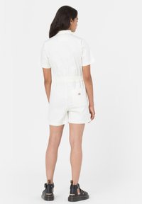 Hvit kort jumpsuit med belte i livet, korte ermer og baklomme. Kombinert med svarte sandaler med stropper. Glatt bomullstekstur.
