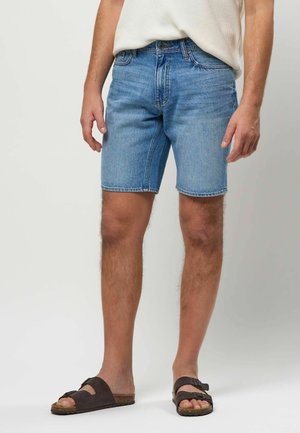 Mężczyzna w jasnoniebieskich dżinsowych szortach, beżowej teksturowanej koszuli i ciemnobrązowych sandałach z klamrą stoi na tle jednolitego tła.