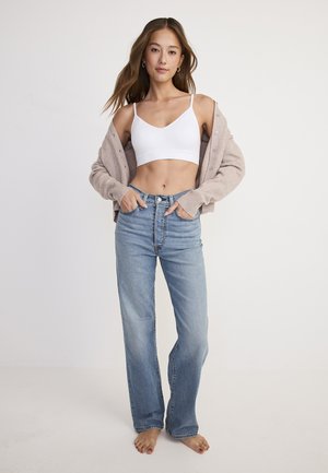 Modelo con un bralette blanco y un cárdigan color beige claro, combinado con jeans azules de corte recto y cintura alta. Sin zapatos. Texturas de tela suaves.