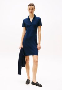 Marineblå polo kjole med korte ærmer, V-hals og tætsiddende silhuet. Accessories inkluderer en vævet marineblå cardigan og sorte loafers.